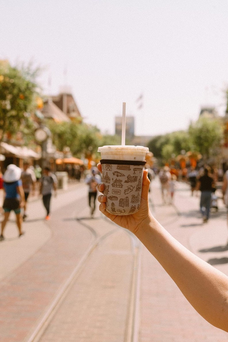 May include: A brown paper cup with a white lid and a straw, filled with a light brown beverage. The cup has a pattern of small black and white illustrations of buildings and other attractions. The cup is being held by a person's hand.