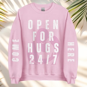 Könnte beinhalten: Ein hellrosa Sweatshirt mit dem Text "OPEN FOR HUGS 24/7" in weißen Blockbuchstaben. Das Wort "COME" ist vertikal auf dem linken Ärmel und "HERE" vertikal auf dem rechten Ärmel aufgedruckt. Eine Grafik von zwei sich umarmenden Figuren befindet sich in der Mitte.