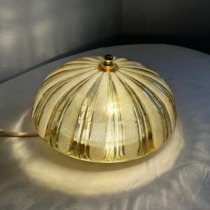 Pode incluir: Uma luminária de mesa decorativa com uma sombra arredondada texturizada de cor dourada. A luminária tem um design canelado e um pequeno remate de cor escura no topo. A luminária está ligada, emitindo um brilho quente.