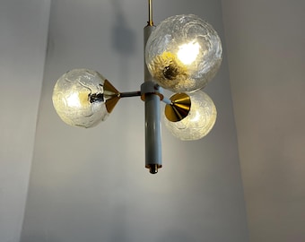Puede incluir: Una lámpara de araña vintage con tres pantallas esféricas de cristal. La lámpara tiene un acabado blanco y dorado, con una columna central y brazos que sostienen las pantallas. Las pantallas de cristal tienen un aspecto texturizado y esmerilado.