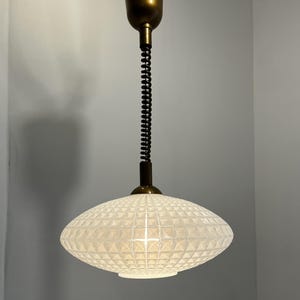 May include: A vintage pendant light with a textured, white glass shade. The shade is a flattened oval shape with a grid pattern. The fixture has a brass-coloured top and a black, coiled support. The light hangs from the ceiling.