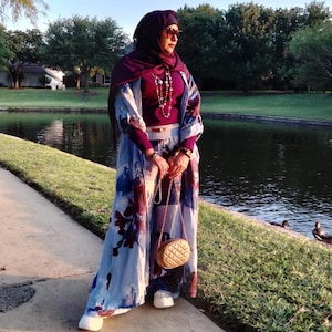 May include: A woman wearing a burgundy top, a floral patterned robe, and matching pants. She is wearing a burgundy hijab and sunglasses. She is holding a small, quilted, tan purse with a gold chain strap. She is standing on a concrete path near a lake.