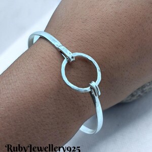 May include: A silver-colored bangle bracelet with a circular clasp. The bracelet is worn on a wrist. The text "RubyJewellery925" is at the bottom of the image.