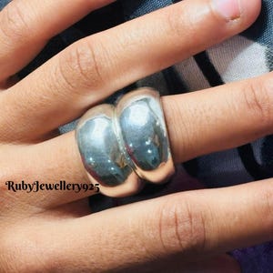 Puede incluir: Un anillo plateado brillante con un diseño único de doble banda. El anillo es liso y pulido, reflejando la luz. El texto "RubyJewellery925" es visible en el lado izquierdo de la imagen. Este anillo es una joya.