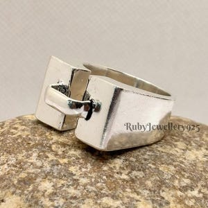 Op de afbeelding: Een zilverkleurige ring met een rechthoekig ontwerp. De ring heeft een uniek gespachtig kenmerk en is geplaatst op een natuurlijk stenen oppervlak. De tekst "RubyJewellery925" is zichtbaar op de ring.