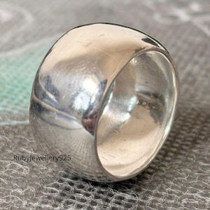 Puede incluir: Un anillo ancho de plata pulida con la parte superior redondeada. El anillo tiene una superficie lisa y reflectante y una banda gruesa. El texto "Rubyjewellery925" es visible en la parte inferior izquierda de la imagen. Una joya llamativa.