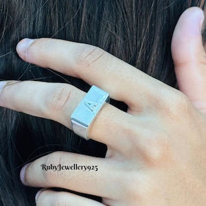 Op de afbeelding: Een zilverkleurige ring met een rechthoekig oppervlak, met de letter "A" in een gestileerd lettertype. De ring wordt aan een vinger gedragen en toont zijn eenvoudige maar elegante ontwerp. De ring is gemaakt van glanzend metaal.