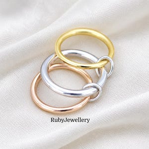 May include: A set of three interlocking rings in yellow gold, silver, and rose gold tones. The rings feature a smooth, polished, and rounded band design. The text RubyJewellery is printed in black at the bottom of the image.