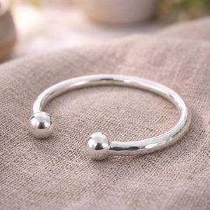 May include: A silver cuff bracelet featuring a polished, hammered texture along the band and two smooth, spherical ball ends. The open-ended design is crafted from a reflective, metallic material with a bright, clean finish and rounded edges.