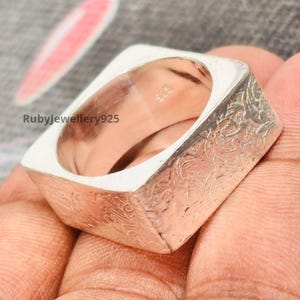 May include: A square silver ring with a textured floral pattern on the sides. The ring has a smooth, polished interior and the text "925" is visible. The ring is shown on a hand.
