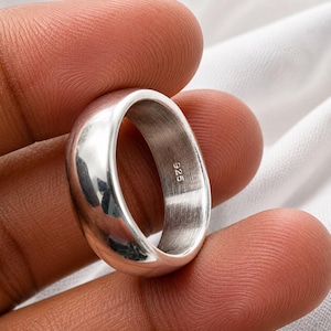 Puede incluir: Un anillo de banda de tono plateado pulido con una superficie exterior lisa y redondeada. El interior del anillo presenta una textura de metal cepillado y está claramente marcado con el texto numérico 925, indicando la pureza del metal.