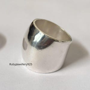Puede incluir: Un anillo de plata con un diseño redondeado y asimétrico. El anillo tiene una superficie lisa y pulida y una banda ancha. El texto "Rubyjewellery925" es visible en el anillo. El anillo es probablemente de plata de ley.