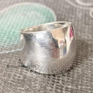 Puede incluir: Un anillo ancho de plata con una superficie texturizada. El anillo tiene una forma redondeada y un acabado pulido, que refleja la luz. El anillo es una joya.