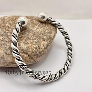 Può includere: Un bracciale a polsino color argento con un design a corda intrecciata. Il bracciale ha una sfera ad ogni estremità. Il bracciale è appoggiato su una roccia grigia. Il testo "RubyJewellery925" è visibile.