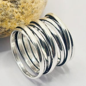 May include: A silver-colored ring with a multi-band design. The ring features multiple, curved bands that wrap around the finger. The bands have a slightly textured appearance, and the ring is set against a white background.