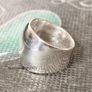 Puede incluir: Un anillo ancho de plata con un diseño liso y curvado. El anillo tiene un acabado pulido y una superficie ligeramente texturizada. El texto "Rubyjewellery925" es visible en el anillo. Esta joya es adecuada para el uso diario.