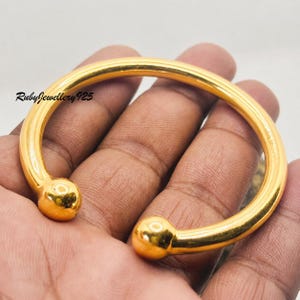 May include: A gold-toned open cuff bracelet with a smooth, polished finish and a spherical ball at each end. The bracelet features a curved, circular design and includes the text RubyJewellery925 printed in black on the left side of the band.