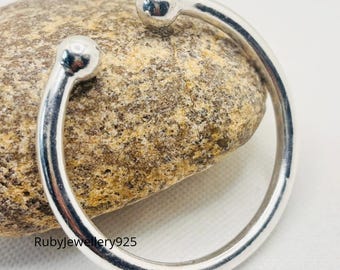 May include: A silver-colored cuff bracelet with a rounded design. The bracelet has a smooth, polished finish and features a small, spherical detail at each end. The bracelet is resting on a textured, light brown stone.