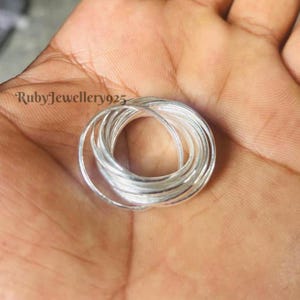 May include: A close-up of a silver ring set, with multiple intertwined bands. The rings are arranged in a circular pattern, reflecting light. The brand name "RubyJewellery925" is visible in the upper left corner. The rings are held in a hand.