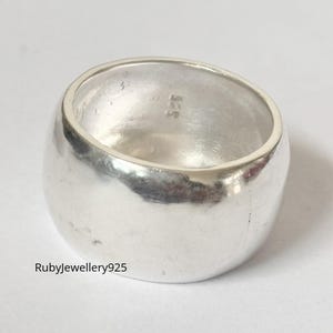 Könnte beinhalten: Ein breiter, polierter Silberring mit abgerundeter Form. Der Ring hat eine glatte, reflektierende Oberfläche und ein schlichtes, minimalistisches Design. Der Ring ist innen mit "925" markiert. Der Ring ist ein Schmuckstück.