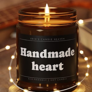 Könnte beinhalten: Eine brennende Kerze in einem braunen Glasgefäß mit schwarzem Etikett, das den Text "ERIN'S CANDLE HEAVEN", "Handmade heart" und "ECO-FRIENDLY | SOY CANDLE" trägt. Das Glas steht auf einem runden Holzuntersetzer, umgeben von einer Lichterkette.