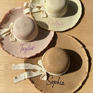 Könnte beinhalten: Vier Sonnenhüte aus Stroh mit breiten Krempen und ausgefransten Rändern. Jeder Hut hat eine runde Krone und eine dekorative Spitzenschleife. Die Hüte sind in Rosa-, Beige- und Brauntönen gehalten und mit den Namen Taylor, Kaitlyn und Sophia bestickt.