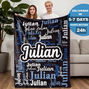 May include: A black blanket with the name "Julian" in white and blue lettering. The name is repeated in various fonts and sizes across the blanket. The blanket is held by two people.