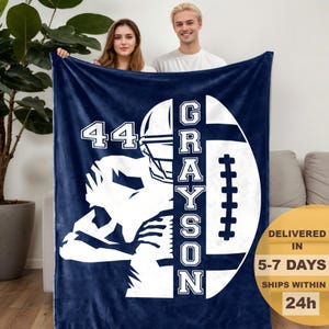 Könnte beinhalten: Marineblaue Fleece-Decke mit weißer Footballspieler-Grafik, der Nummer 44 und dem Namen "Grayson" in vertikaler Schrift. Die Decke hat eine Footballform. Die Decke wird von zwei Personen gehalten.