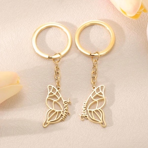 Pode incluir: Dois chaveiros dourados com borboletas. Cada chaveiro apresenta um design de asa de borboleta com um padrão vazado. Uma asa é inscrita com o nome "Evelyn" e a outra com "Fiona". Os chaveiros têm um anel e uma corrente dourados.