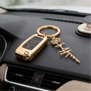 May include: A gold-colored car key with a matching key ring and a decorative charm. The charm features a flower and the word "Mom" in a stylized script. The key and charm rest on a black dashboard.