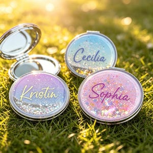 Könnte beinhalten: Silberne Kompaktspiegel mit den Namen "Cecilia", "Kristin" und "Sophia". Jeder Spiegel hat ein farbenfrohes, glitzergefülltes Design. Ein Spiegel ist geöffnet und zeigt die reflektierende Oberfläche. Auf grünem Gras platziert.