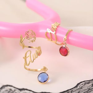 May include: Gold-colored rings with personalized names and gemstone accents. One ring features a flower design, while others have a name inscription and a dangling gemstone. The gemstones are red and blue.