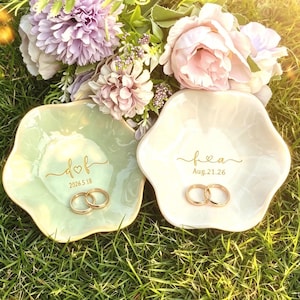 May include: Two ceramic ring dishes, one pale green and one white, each holding two gold wedding rings. The green dish has the inscription "dof" and the date "2026.5.18". The white dish has the inscription "fea" and the date "Aug.21.26".