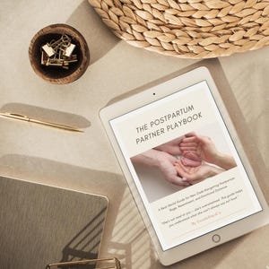 May include: A digital tablet shows "The Postpartum Partner Playbook" with a photo of hands holding a baby's feet. A gold pen, a small wooden bowl with gold paper clips, and a woven basket are also visible.