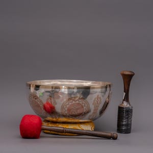 Puede incluir: Un cuenco tibetano plateado con ribetes dorados y diseños decorativos, acompañado de una maza de madera con un percutor rojo y un percutor de madera. El cuenco descansa sobre un cojín amarillo.