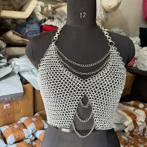 以下が含まれることがあります： 黒いマネキンに展示されたシルバーのチェーンメイルクロップトップ。トップはホルターネックのデザインと一連のぶら下がっているチェーンが特徴です。チェーンメイルは、小さな相互接続された金属リングでできており、メッシュのような外観になっています。