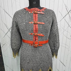 Peut inclure: Une cotte de mailles avec des détails en cuir rouge et une ceinture. La chemise est faite d'anneaux métalliques et a une bordure dorée aux manches et à l'ourlet. Les sangles en cuir rouge sont fixées par des boucles en laiton.