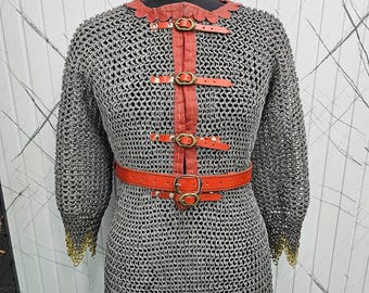 May include: A chainmail shirt with red leather detailing and a belt. The shirt is made of metal rings and has a gold trim at the sleeves and hem. The red leather straps are secured with brass buckles.