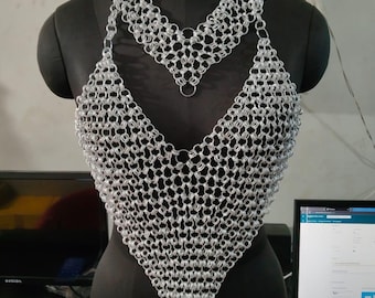 May include: A silver chainmail top and necklace displayed on a black mannequin. The chainmail is made of small, interlocking metal rings, forming a V-neck top and a matching choker-style necklace. The top is sleeveless.