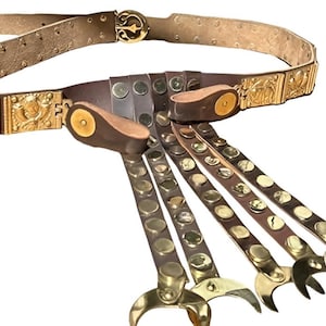 May include: A brown leather belt with gold-colored metal accents. The belt features a decorative buckle and ornate rectangular plates. Several leather straps with gold studs and claw-shaped ends hang from the belt.