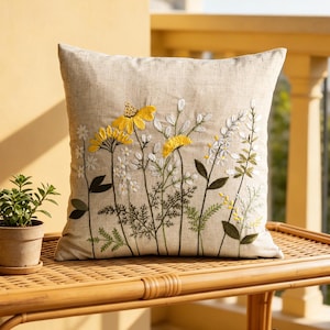 May include: A square beige linen throw pillow featuring detailed embroidery of yellow and white wildflowers with green stems and leaves. The floral design is centered on the front of the pillow, showcasing various botanical shapes and textures.