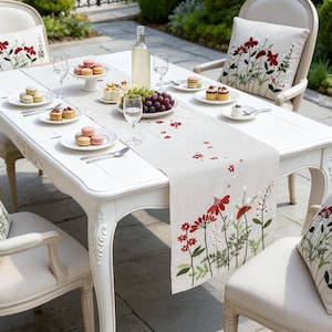 Op de afbeelding: Een witte houten eettafel met vier bijpassende stoelen met crèmekleurige bekleding. Elke stoel is voorzien van een vierkant kussen met rode bloemenborduursels. Een linnen tafelloper met bijpassende rode bloemmotieven ligt in het midden.