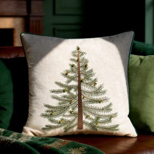 Può includere: Un cuscino quadrato con una fodera color beige chiaro effetto lino, caratterizzato da un ricamo centrale raffigurante un pino verde. L'albero presenta un tronco marrone e piccoli ornamenti rotondi in metallo dorato tra i rami.