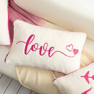 May include: White rectangular pillow with the word "love" embroidered in bright pink script, accompanied by a small heart. The pillow has a pink border and is on a white surface.