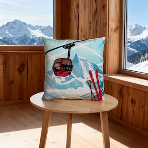 Puede incluir: Un cojín cuadrado con un diseño de acuarela que presenta un teleférico rojo, esquís rojos y una escena de montaña en tonos azules y blancos. El cojín está sobre una mesa de madera marrón claro.