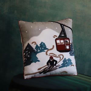 Può includere: Cuscino quadrato decorativo con una scena di stazione sciistica. Il cuscino presenta uno sciatore, una funivia rossa e alberi sempreverdi ricamati su uno sfondo bianco. Il cuscino ha una parte superiore grigia con neve e un bordo rosso.