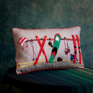 Op de afbeelding: Een rechthoekig kussen met een lichtgrijze achtergrond en een rode rand. Het kussen heeft geborduurde ski-uitrusting, waaronder rode ski's, een groen snowboard, een rood-witte muts en bruine wanten. Het ontwerp roept een wintersportthema op.