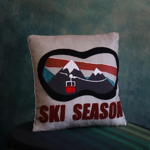 Puede incluir: Un cojín decorativo cuadrado de color gris claro con un diseño de gafas de esquí. Las gafas enmarcan una escena de montaña con picos nevados, un telesilla y las palabras "SKI SEASON" en rojo.