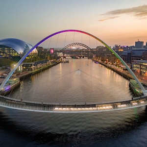 Può includere: Vista aerea al tramonto di un moderno ponte ad arco multicolore che attraversa un fiume. Il ponte è circondato da edifici e altri ponti. L'acqua riflette le luci della città, creando un'atmosfera notturna.