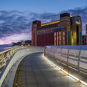 Puede incluir: Una vista nocturna de Baltic Flour Mill, un edificio de ladrillo con el texto "BALTIC FLOUR MILL" en su fachada. Un puente peatonal curvo con luces conduce al edificio bajo un cielo nublado.